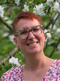 Eine Person mit Brille lächelt vor blühenden weißen Blumen im Hintergrund.