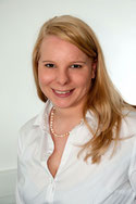 Eine lächelnde Person mit langen blonden Haaren trägt eine weiße Bluse vor einem neutralen Hintergrund.