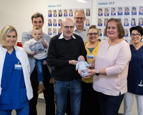 Eine Gruppe von sieben Erwachsenen und einem Kleinkind posiert lächelnd in einem Krankenhausflur.