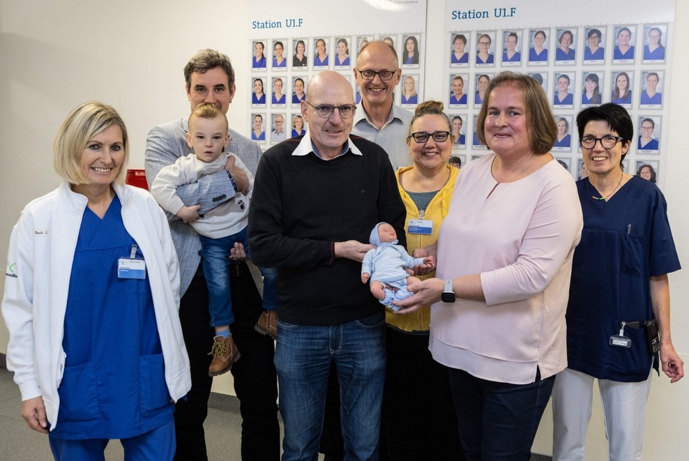 Eine Gruppe von sieben Erwachsenen und einem Kleinkind posiert lächelnd in einem Krankenhausflur.