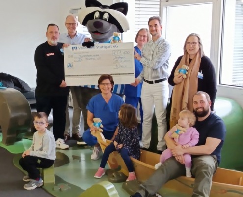 Eine Gruppe von Erwachsenen und Kindern hält einen großen Scheck in einem Raum mit Spielzeug. Ein Maskottchen ist dabei.