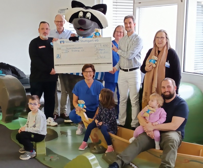 Eine Gruppe von Erwachsenen und Kindern hält einen großen Scheck in einem Raum mit Spielzeug. Ein Maskottchen ist dabei.