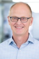 Ein lächelnder Mann mit Brille und blauem Hemd vor unscharfem Hintergrund.