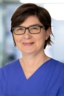 Eine Person mit Brille und blauer Kleidung lächelt vor unscharfem Hintergrund.