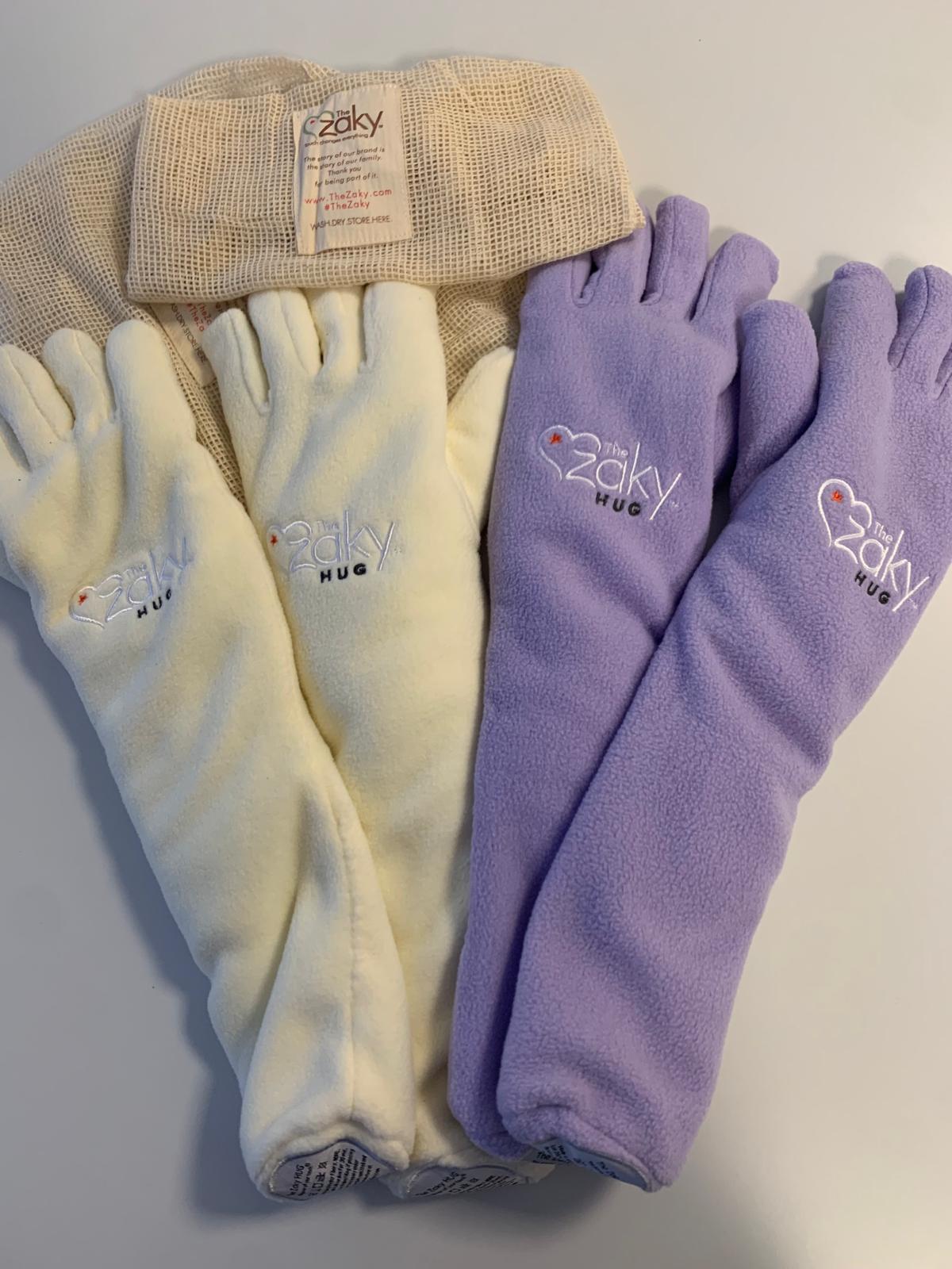 Zwei cremefarbene und zwei lila Babyhandschuhe liegen auf einem Tisch neben einem beigen Netzbeutel.