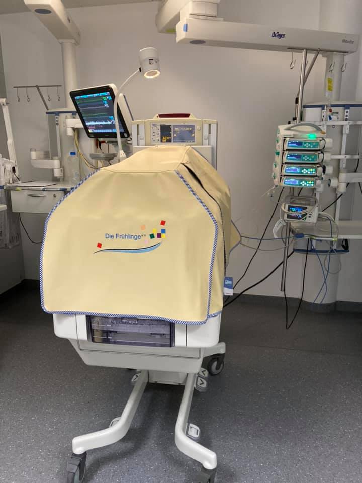 Ein Inkubator mit gelber Abdeckung und der Aufschrift Die Frühlinge e.V. in einem Krankenhausraum, umgeben von medizinischen Geräten und Monitoren.